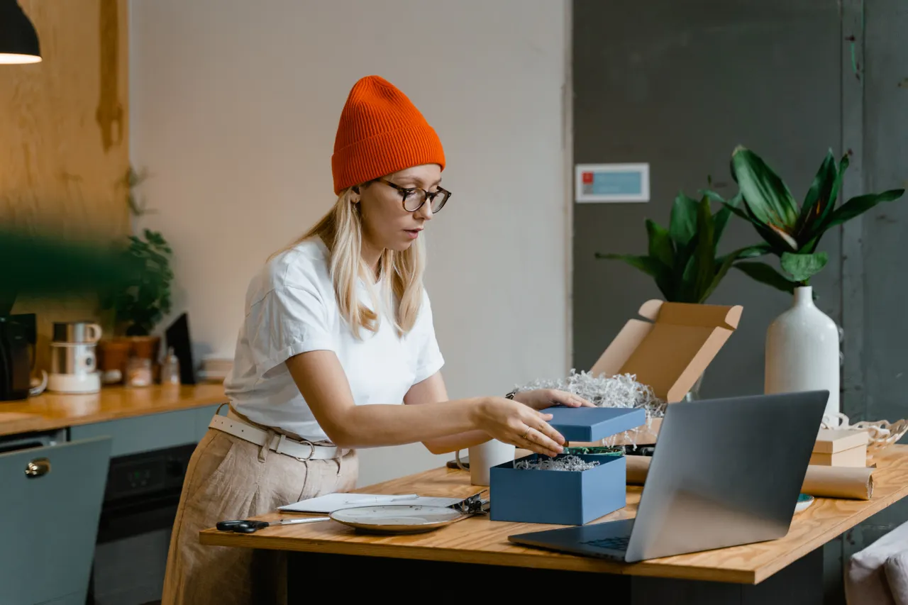 Une commerçante prépare un colis dans une boîte bleue près de son ordinateur portable.